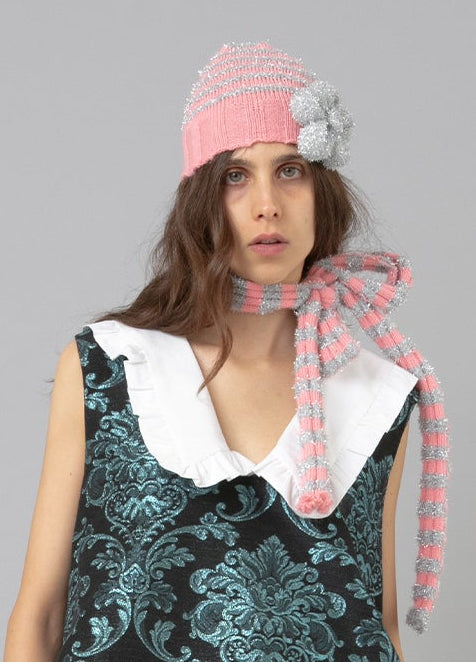 A model stands against a solid gray studio background wearing a sleeveless black and teal damask-patterned pinafore dress with a white collar, gray knee-high socks, black shoes, and a pink striped knit hat with a bow.