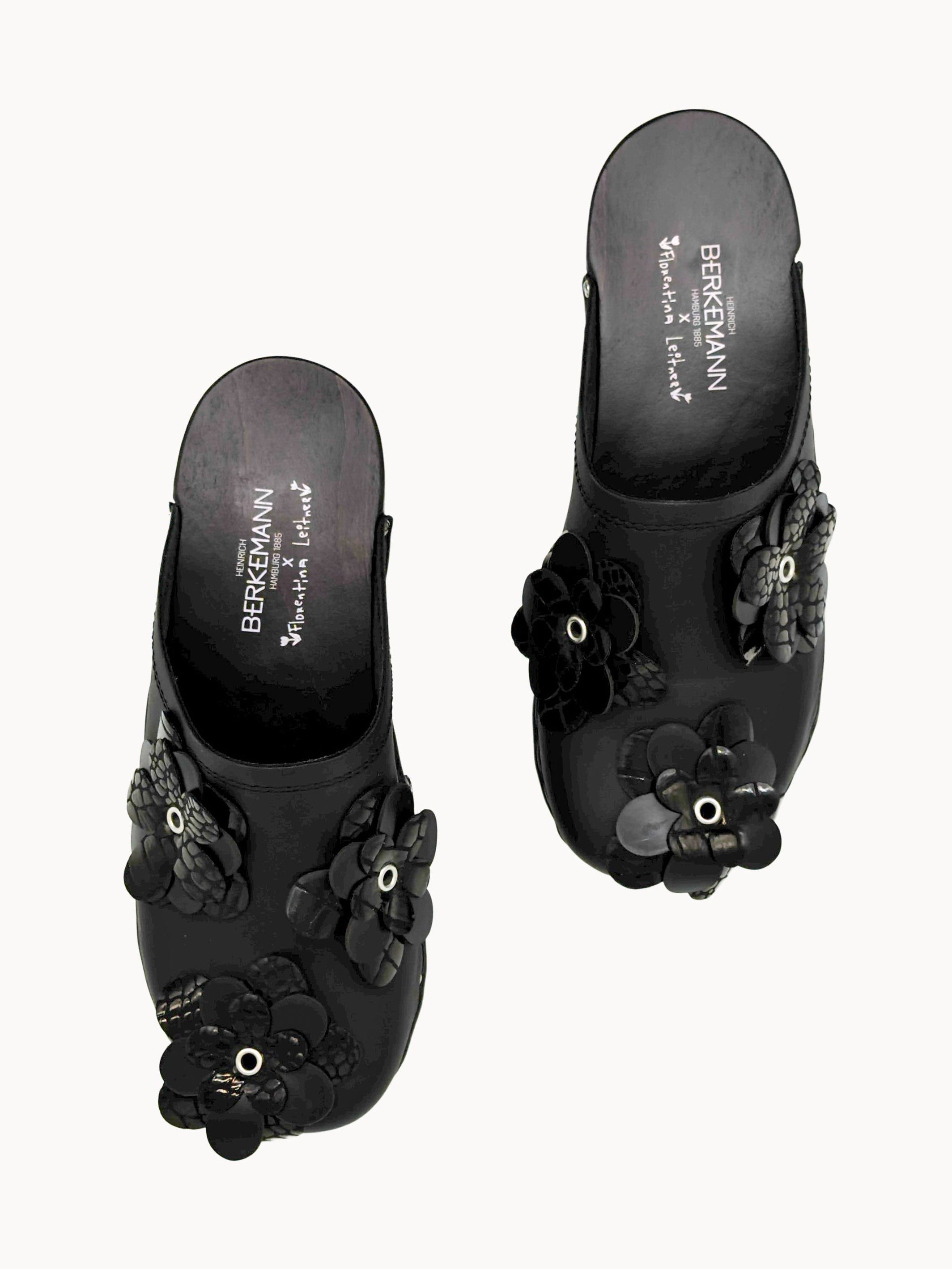A pair of black slide sandals with 3D floral embellishments and rhinestone details, shown from a top-down view on a light gray background.